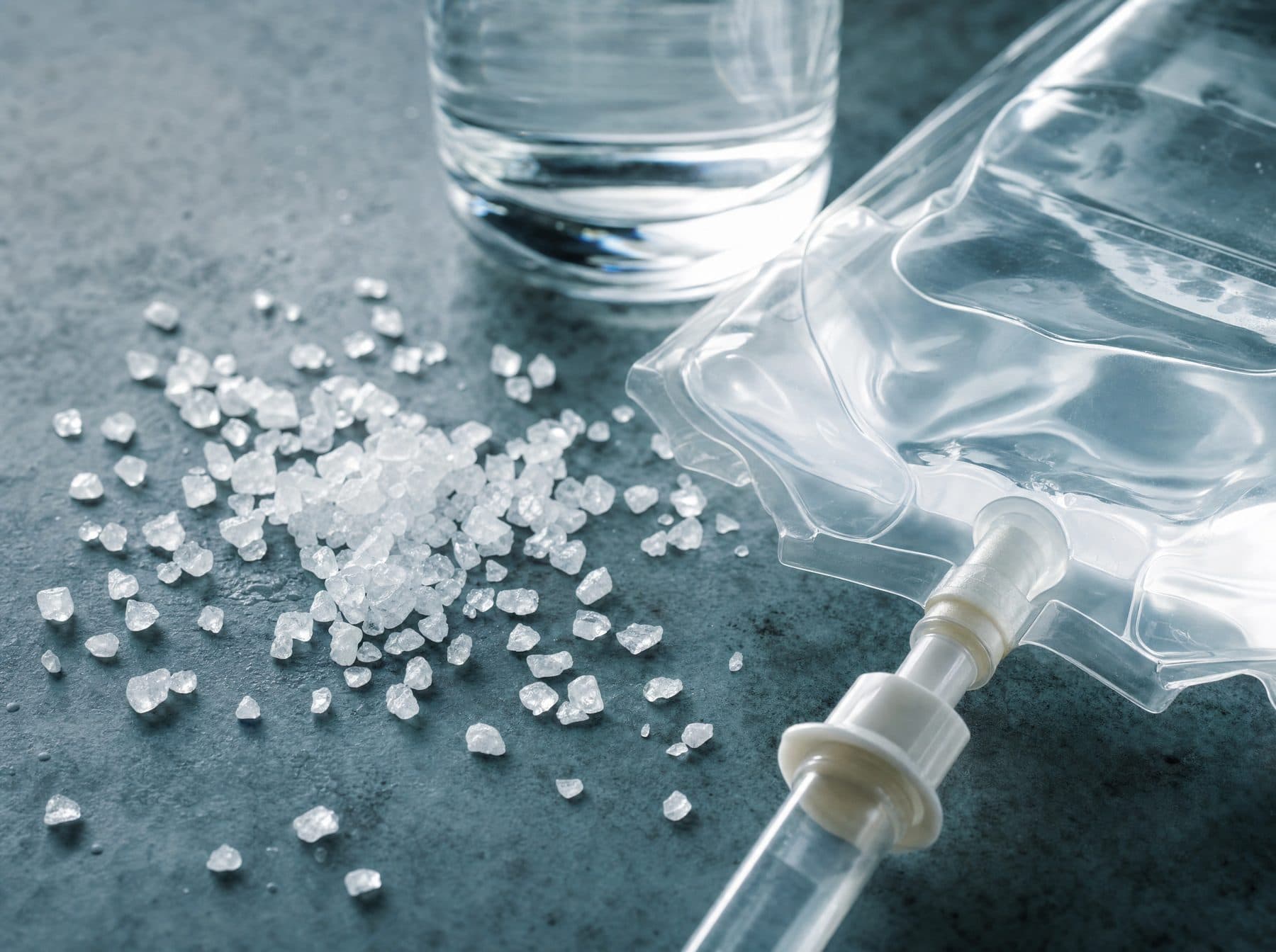 Salt crystals beside a clear saline IV bag and a glass of water